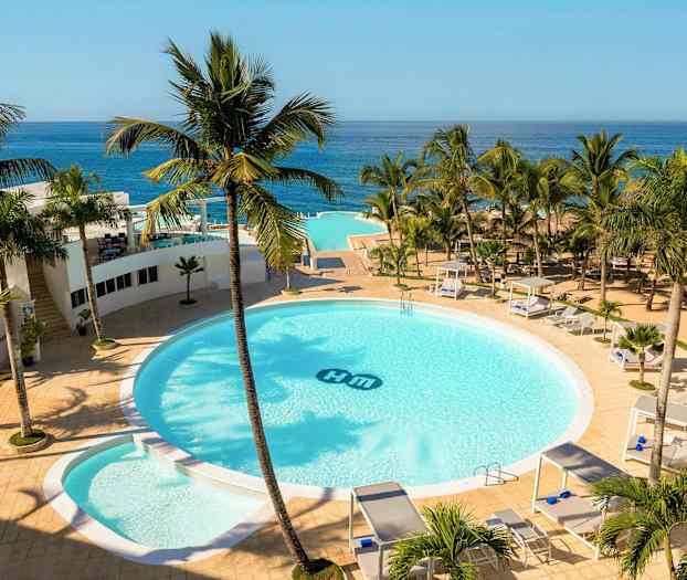 Hotellialue, jossa on suuri pyöreä uima-allas, ympäröity palmuilla, aurinkotuoleilla ja cabanoilla. Taustalla näkyy merenranta. (Tekoälyn luoma teksti)