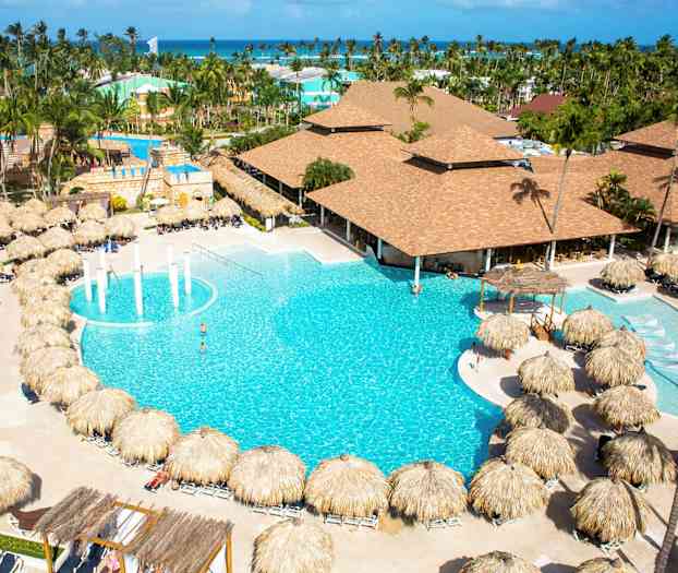 Hotellialue, jossa on suuri uima-allas, aurinkotuoleja palmakattoineen ja taustalla rakennuksia. (Tekoälyn luoma teksti)