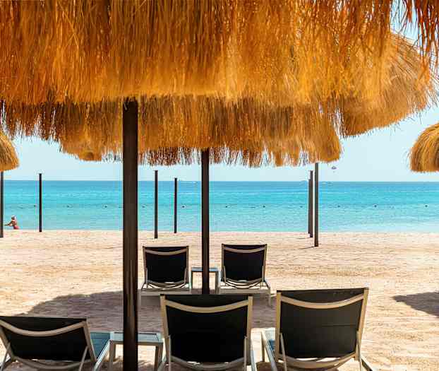 Hiekkaranta, jolla aurinkotuoleja ja olkikattoisia varjoja, sekä meri taustalla. (Tekoälyn luoma teksti)