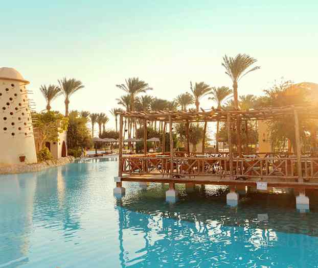 Kuva uima-altaasta, jossa on puinen paviljonki ja pieniä reikiä sisältävä rakennus palmujen ympäröimänä. (Tekoälyn luoma teksti)