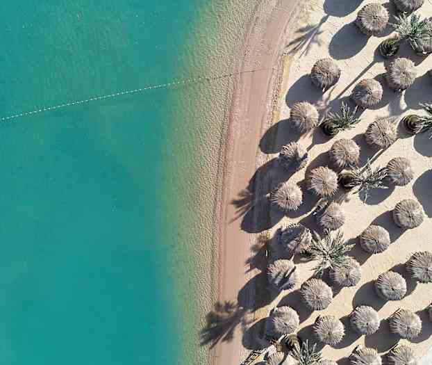 Ilmakuva hiekkarannasta, jossa turkoosi vesi ja aurinkovarjoja. (Tekoälyn luoma teksti)