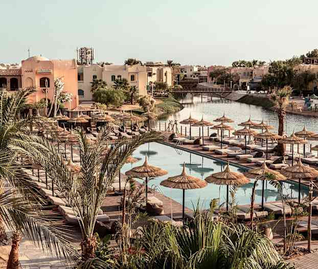 Uima-allasalue, jossa aurinkotuoleja ja aurinkovarjoja, ympäröity palmuilla ja rakennuksilla. (Tekoälyn luoma teksti)