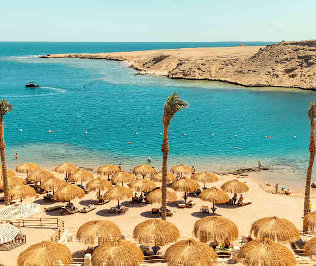 Hiekkaranta varjoilla, palmuilla ja ihmisillä veden äärellä. (Tekoälyn luoma teksti)