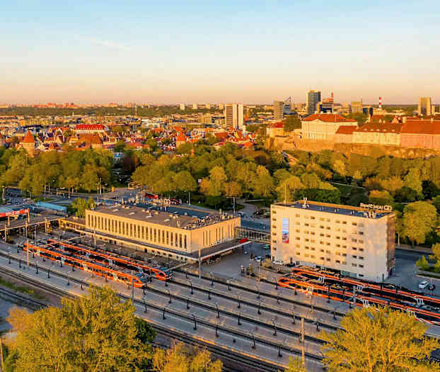 Kuva kaupungista, jonka etualalla on rautatieasema ja taustalla kaupungin siluetti rakennuksineen ja puineen. (Tekoälyn luoma teksti)