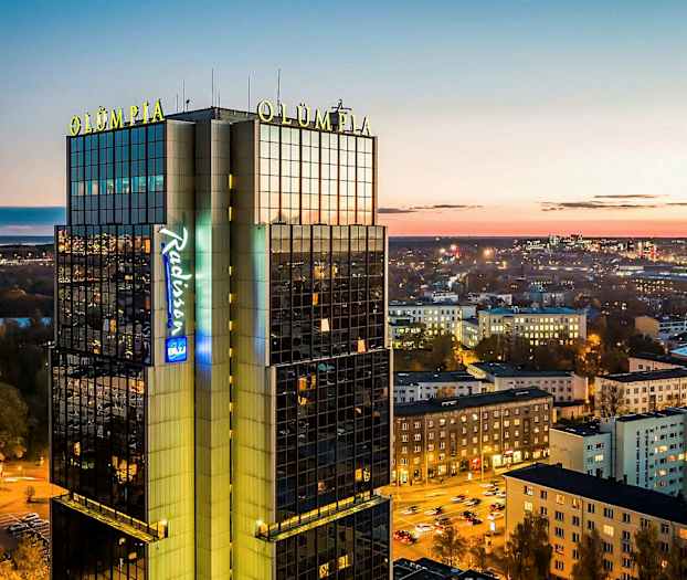 Korkea rakennus, jossa lukee "Olümpia" ja "Radisson", kaupunkinäkymä hämärässä. (Tekoälyn luoma teksti)