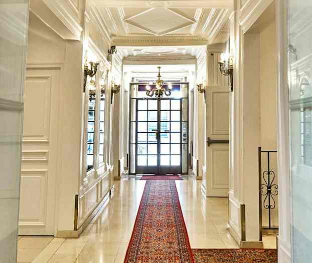 Tyylikäs käytävä, jossa on valkoiset seinät ja lattia, punainen matto sekä kattokruunu ja lasiovet taustalla. (Tekoälyn luoma teksti)