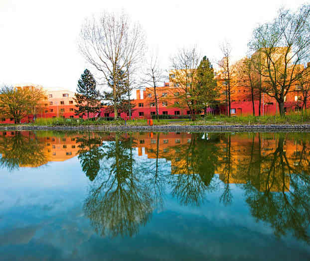 Kuva punaisista rakennuksista, joita ympäröivät puut ja jotka heijastuvat tyynestä vedenpinnasta. (Tekoälyn luoma teksti)