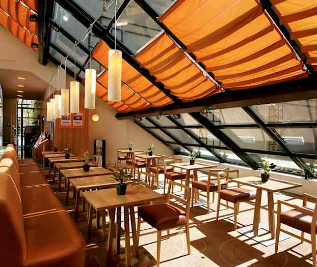 Valoisa ja moderni ruokailutila, jossa on puupöytiä ja tuoleja, oranssit kattokoristeet ja suuret ikkunat. (Tekoälyn luoma teksti)