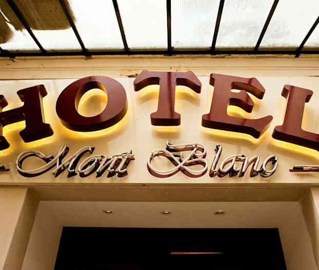 Hotellin kyltti, jossa lukee "HOTEL Mont Blanc"; "HOTEL" on suurilla, valaistuilla kirjaimilla ja "Mont Blanc" on kursiivilla koristeellisessa tyylissä. (Tekoälyn luoma teksti)