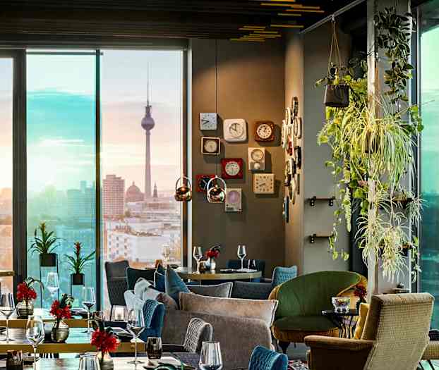 Tyylikäs ravintolan sisustus, jossa on modernit kalusteet, vihreitä kasveja ja näkymä kaupungin siluettiin tunnusrakennuksen kanssa. (Tekoälyn luoma teksti)
