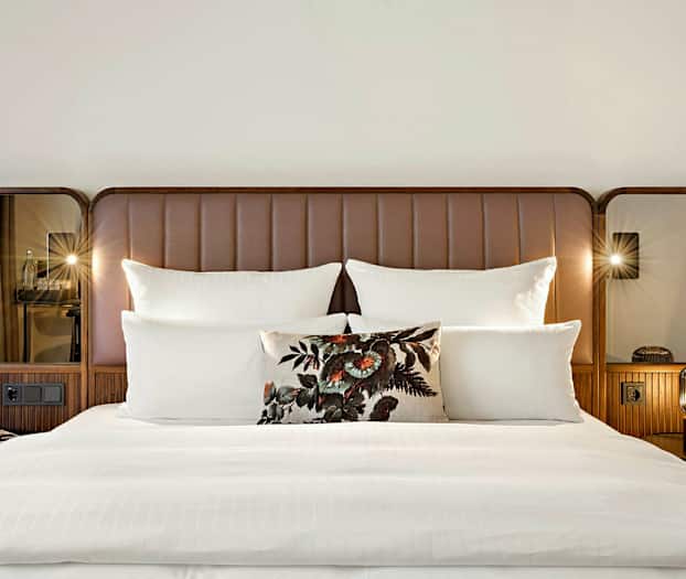 Parivuode, jossa on valkoinen vuodevaatteet, useita tyynyjä ja koristeellinen kukkakuvioinen tyyny. Sänkyyn liittyvä pääty on ruskea ja siinä on peilit sivuilla. (Tekoälyn luoma teksti)