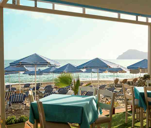 Ranta, jossa sinivalkoraitaisia aurinkovarjoja ja aurinkotuoleja, pöytiä etualalla. (Tekoälyn luoma teksti)