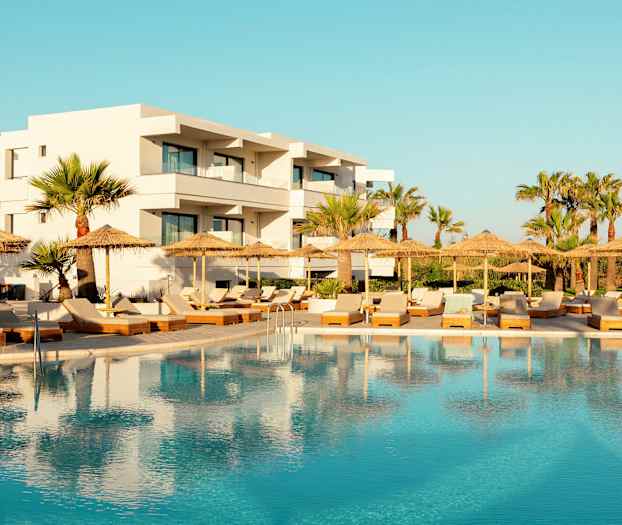 Hotelli valkoisella julkisivulla uima-altaan ja aurinkotuolien takana, joissa on aurinkovarjoja. (Tekoälyn luoma teksti)