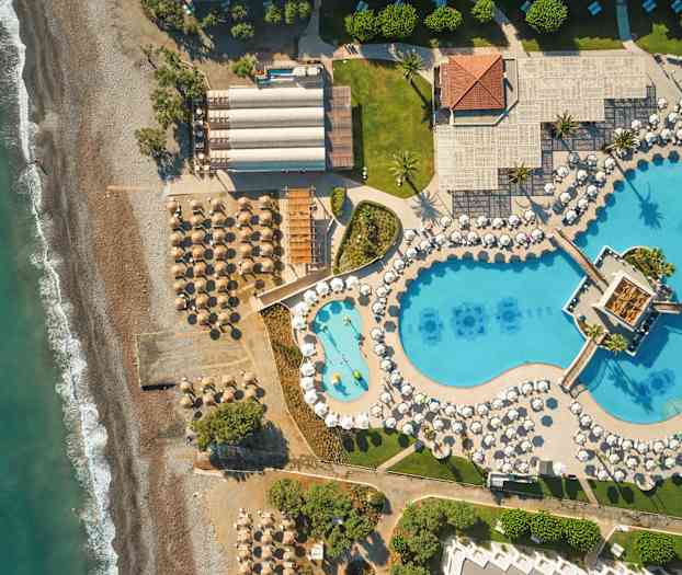 Ilmakuva rannasta, jolla on aurinkovarjoja ja aurinkotuoleja, sekä vieressä suuri allasalue, jossa sinisiä uima-altaita ja valkoisia aurinkovarjoja. (Tekoälyn luoma teksti)