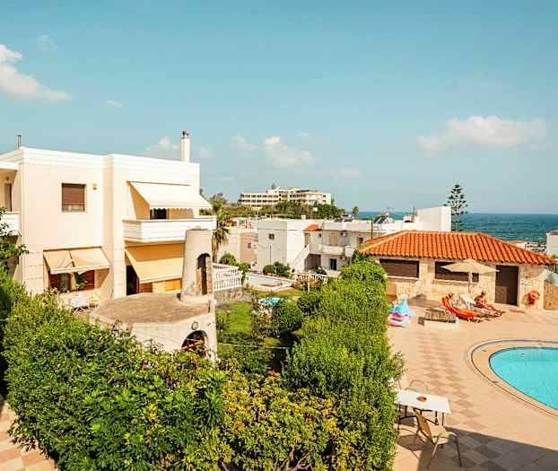 Kuvassa näkyy ulkoalue, jossa on taloja, vihreitä kasveja, sininen uima-allas ja aurinkotuoleja aurinkoisena päivänä. (Tekoälyn luoma teksti)