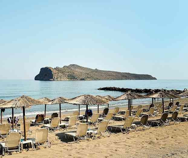 Aurinkotuoleja ja varjoja hiekkarannalla, taustalla kallio meressä. (Tekoälyn luoma teksti)