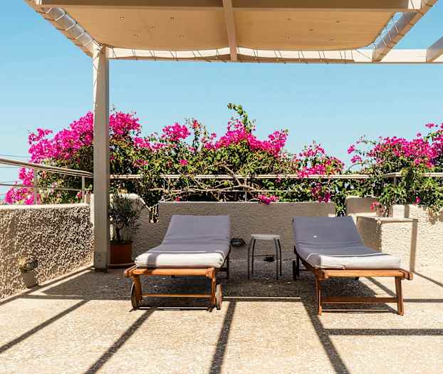 Terassi, jossa on kaksi aurinkotuolia, bougainvillea ja katos sinisen taivaan alla. (Tekoälyn luoma teksti)