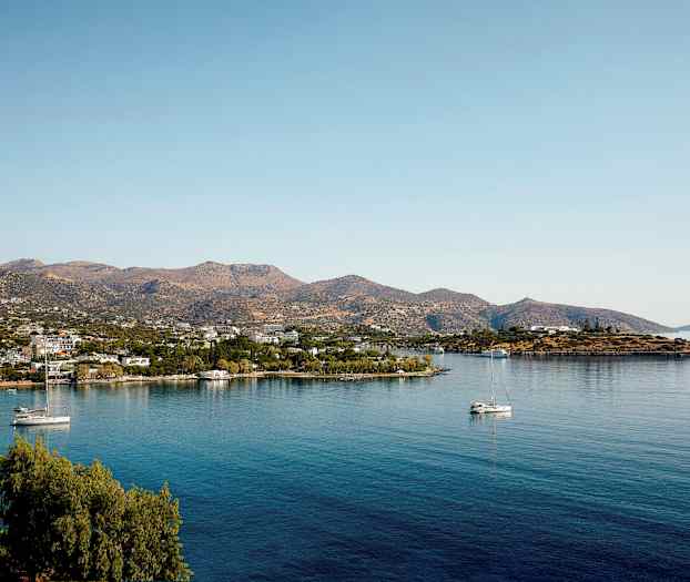 Maisema rauhallisella lahdella, purjeveneitä ja pieniä rakennuksia vuoren juurella. (Tekoälyn luoma teksti)