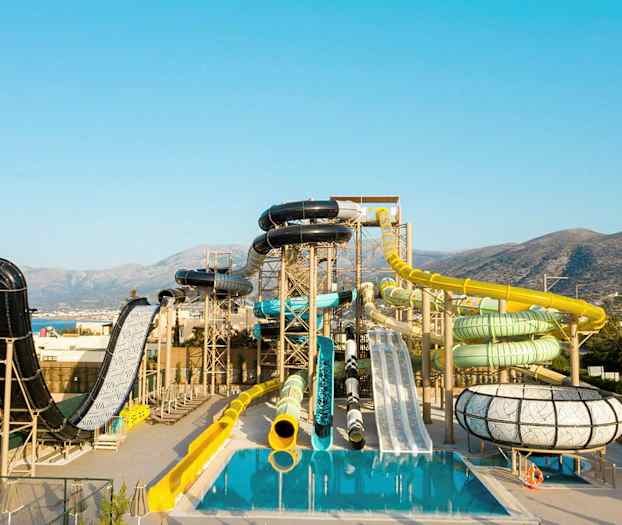 Vesipuisto, jossa on erilaisia liukumäkiä ja altaita vuoristoisen taustan edessä. (Tekoälyn luoma teksti)