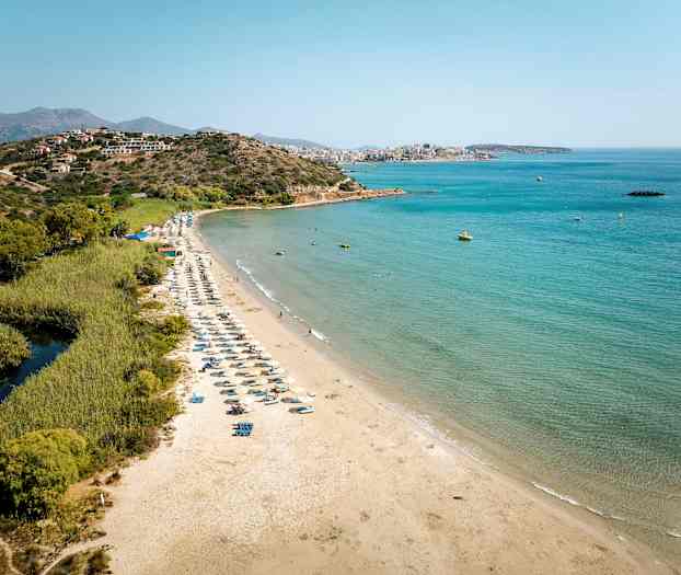Hiekkaranta aurinkotuoleineen meren äärellä, ympärillä vehreyttä ja taustalla vuoria. (Tekoälyn luoma teksti)