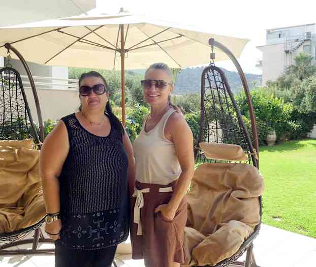 Kaksi riippukeinua suuren aurinkovarjon alla terassilla, josta on näkymä puutarhaan ja ympäröiviin rakennuksiin. (Tekoälyn luoma teksti)