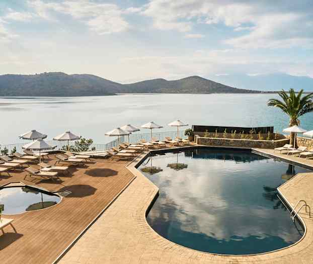 Uima-allas aurinkotuoleilla ja varjoilla meren äärellä, ympäröitynä vuorilla ja palmuilla. (Tekoälyn luoma teksti)