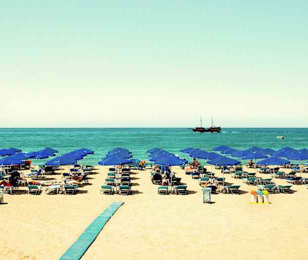 Ranta, jossa sinisiä aurinkovarjoja, aurinkotuoleja ja laiva merellä. (Tekoälyn luoma teksti)