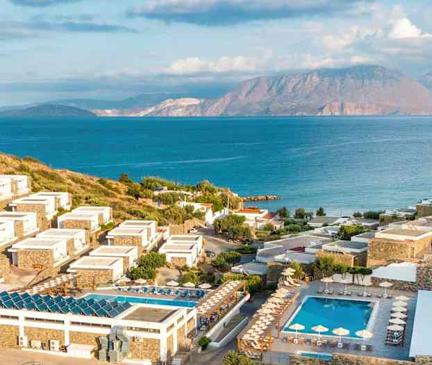 Kuva rannikon lomakeskuksesta, jossa hotellirakennuksia rinteessä, ympärillä vihreitä alueita ja uima-altaita, taustalla meri ja vuoret. (Tekoälyn luoma teksti)