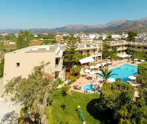 Ilmakuva hotellista, jossa on uima-allas ja puutarha, ympäröitynä vehreydellä ja rakennuksilla, taustalla vuoria. (Tekoälyn luoma teksti)