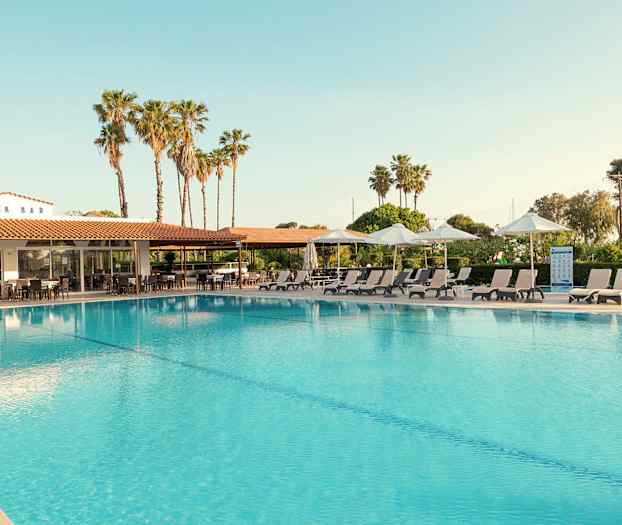 Ulkouima-allas, jonka reunalla on aurinkotuoleja ja aurinkovarjoja. Taustalla näkyy rakennus, jossa on kyltti 'SNACK BAR', ja palmuja. (Tekoälyn luoma teksti)