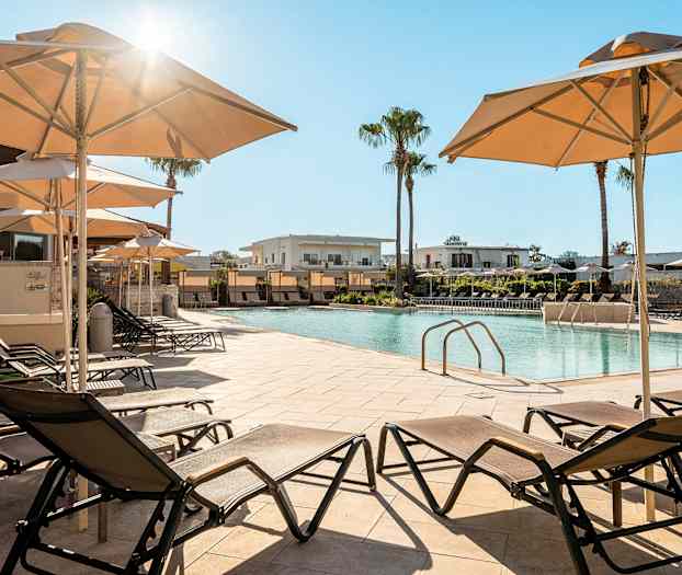 Kuva uima-allasalueesta, jossa on aurinkotuoleja ja aurinkovarjoja, taustalla rakennuksia. (Tekoälyn luoma teksti)