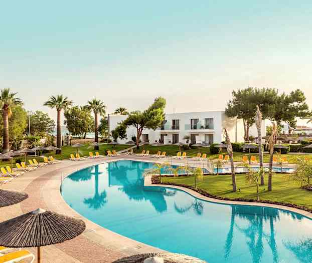 Suuri uima-allas, jonka ympärillä on aurinkotuoleja ja aurinkovarjoja, ja taustalla vihreitä nurmikoita sekä palmuja ja valkoisia rakennuksia. (Tekoälyn luoma teksti)