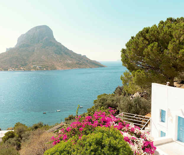 Valkoinen rakennus, jossa siniset ovet, ympäröitynä vihreällä kasvillisuudella ja vaaleanpunaisilla kukilla, näkymänä meri ja vuori taustalla. (Tekoälyn luoma teksti)