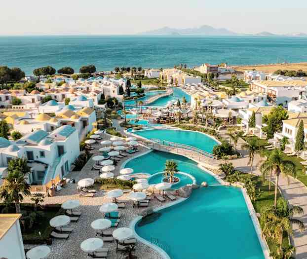 Näköala lomakeskukseen, jossa on uima-altaita, valkoisia rakennuksia ja näkymä merelle taustalla. (Tekoälyn luoma teksti)