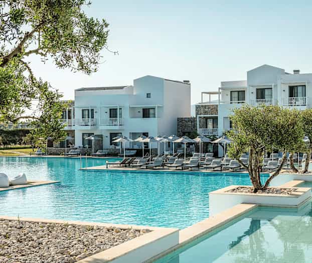 Kuvassa näkyy hotelli, jonka julkisivu on valkoinen, suuren uima-altaan äärellä; ympärillä aurinkotuoleja ja vihreää kasvillisuutta. (Tekoälyn luoma teksti)