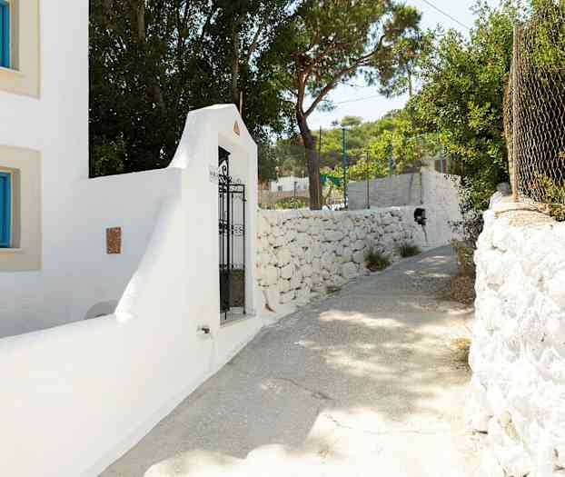 Valkoinen rakennus sinisillä ikkunankehyksillä ja rautaportti kivimuurin luona. Aurinkoinen polku puiden ympäröimänä. (Tekoälyn luoma teksti)