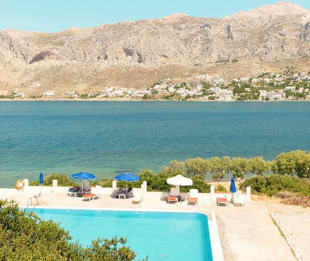 Näkymä uima-altaaseen, jossa on aurinkotuoleja ja varjoja, taustalla meri ja vuoret. (Tekoälyn luoma teksti)