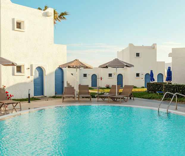 Uima-allas, jota ympäröivät aurinkotuolit ja varjot, taustalla valkoisia rakennuksia sinisillä ovilla. (Tekoälyn luoma teksti)