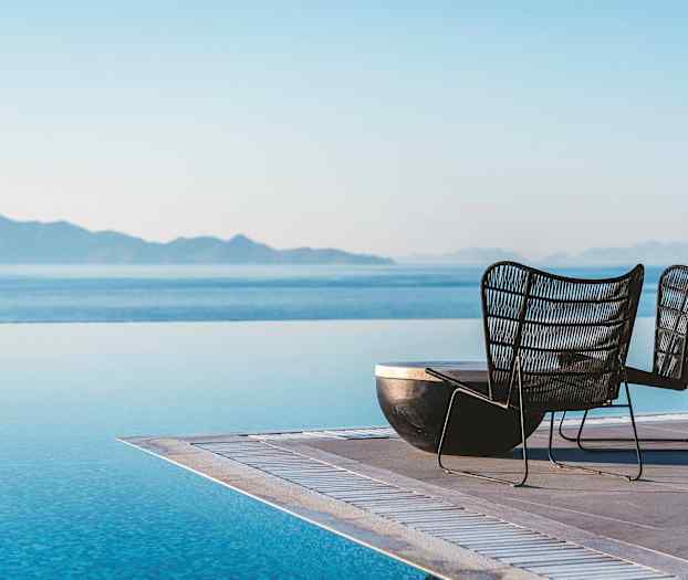 Kaksi tuolia infinity-allasreunalla, taustalla meri ja vuoret. (Tekoälyn luoma teksti)
