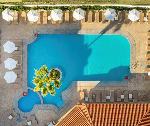 Lintu näkymä suorakaiteen muotoisesta uima-altaasta, jonka ympärillä on aurinkotuoleja ja varjoja. (Tekoälyn luoma teksti)