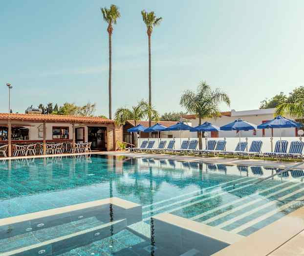 Uima-allas, jota ympäröivät aurinkotuolit ja aurinkovarjot, taustalla baari. (Tekoälyn luoma teksti)