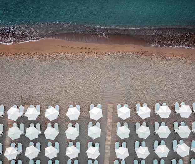 Lintuperspektiivi rannasta, valkoisia aurinkovarjoja ja aurinkotuoleja merenrannassa. (Tekoälyn luoma teksti)