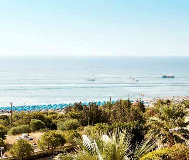 Ranta, jossa sinisiä aurinkovarjoja, ja taustalla meri. Useita veneitä näkyy vedessä. (Tekoälyn luoma teksti)