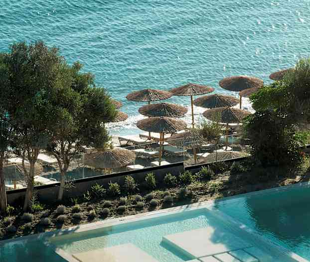 Näkymä altaalle, puille ja rannalle, jossa on aurinkovarjoja ja lepotuoleja kohti merta. (Tekoälyn luoma teksti)