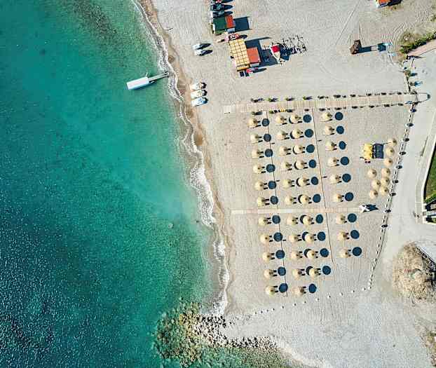 Ilmasta otettu kuva rannasta, jossa on aurinkovarjoja ja aurinkotuoleja kirkkaan sinisen veden vieressä. (Tekoälyn luoma teksti)