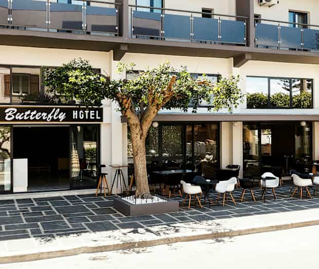 Hotellin etupuoli, jossa on "Butterfly Hotel" -kyltti, ulkoterassi pöytineen ja tuoleineen sekä puu rakennuksen edessä. (Tekoälyn luoma teksti)