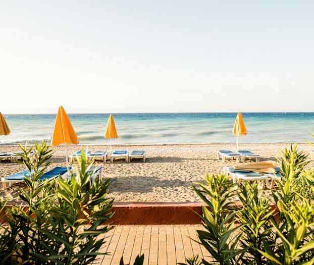 Ranta, jossa aurinkotuoleja ja keltaisia varjoja sinisen meren äärellä, kasvien ympäröimänä. (Tekoälyn luoma teksti)