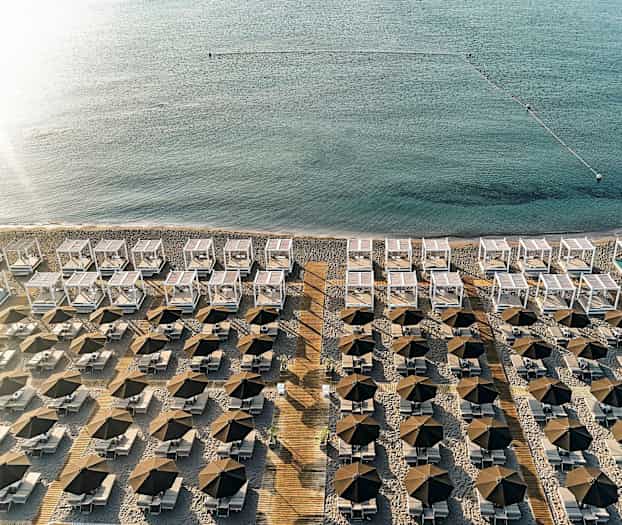 Näkymä rannalta, jolla on riveissä aurinkovarjoja, aurinkotuoleja ja valkoisia aurinkosuojia lähellä merta. (Tekoälyn luoma teksti)