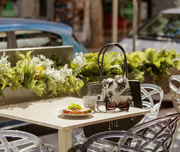 Pöytä, jossa croissant, kuppi ja aurinkolasit ulkoilmaterassilla kukkakoristeilla. (Tekoälyn luoma teksti)