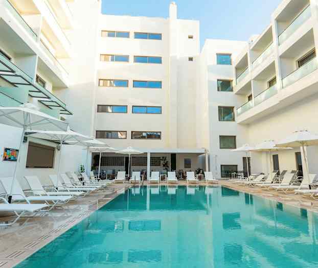 Kuva hotellialueesta, jossa uima-allas on ympäröity aurinkotuoleilla ja varjoilla. Taustalla näkyy valkoisia parvekkeellisia rakennuksia. (Tekoälyn luoma teksti)